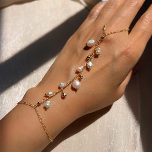 Könnte beinhalten: Ein goldenes Kettenarmband mit einem zarten Perlenmuster. Das Armband hat einen Ring, der am Finger getragen werden kann, und die Kette verläuft mit mehreren Perlenakzenten am Handgelenk entlang.
