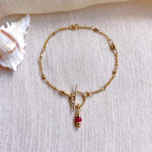 Op de afbeelding: Een delicate goudkleurige armband met een tuimelsluiting en een kleine, hangende hanger. De hanger heeft een cluster van kleine, ronde, rode kralen. De armband wordt gepresenteerd op een crèmekleurige stof.