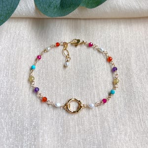 Könnte beinhalten: Goldfarbenes Armband mit einer Mischung aus bunten Perlen und kleinen Perlen. Das Armband hat einen kreisförmigen Goldanhänger und eine zarte Kette. Die Perlen sind in verschiedenen Farben gehalten, darunter Rot, Rosa, Blau, Gelb und Lila.