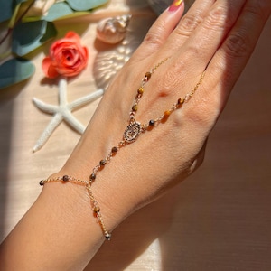 Può includere: Bracciale a catena per mano dorato con un medaglione centrale e piccole perle rotonde marroni e dorate. Il bracciale è collegato a un anello. Lo sfondo include una stella marina e una rosa.