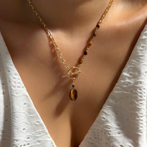 May include: Gold-toned necklace featuring a toggle clasp and a pendant with a tiger's eye stone. The necklace has a chain design with a series of small, round beads in shades of brown and gold. The necklace is worn on a person.