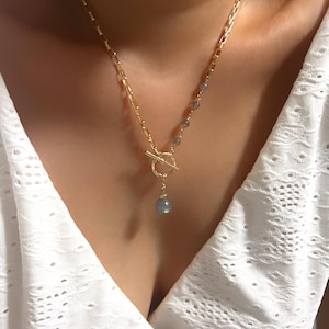 Op de afbeelding: Een goudkleurige ketting met een tuimelsluiting en een hangende blauwgrijze edelsteen hanger. De ketting heeft een kettingontwerp met kleine, ronde edelstenen verspreid langs de ketting.