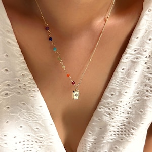 May include: A delicate gold necklace featuring a rainbow of colorful beads and a small rectangular pendant with a starburst design. The necklace is worn against a white eyelet fabric.