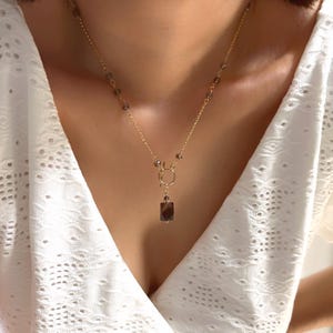 May include: A gold-coloured necklace with a pendant featuring a rectangular, dark brown gemstone. The necklace has a delicate chain with small, dark brown beads and a circular accent near the pendant. The necklace is worn against a white top.