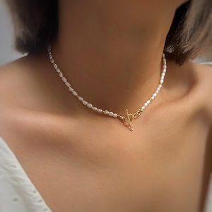 May include: A necklace featuring small, white, irregularly shaped freshwater pearls strung together with tiny gold-toned beads, finished with a gold-toned metal toggle clasp closure positioned at the front center of the neck.