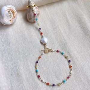 Könnte beinhalten: Ein goldfarbenes Handkettenarmband mit einer filigranen Kette, bunten Perlen und einer großen weißen Perle. Die Kette verbindet einen Ring und ein Armband und schafft ein einzigartiges Schmuckstück.