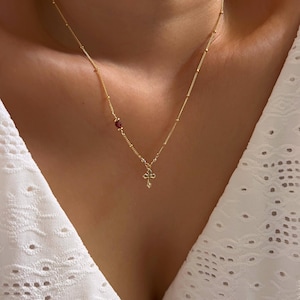 Peut inclure: Un délicat collier en or avec un petit pendentif en forme de croix et une seule pierre rouge. Le collier est porté sur un fond blanc, soulignant le design complexe et le contraste de couleurs du bijou. La chaîne mesure environ 45 cm.