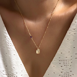 May include: A gold chain necklace with a small rectangular pendant and a single purple gemstone. The necklace is on a person's neck.
