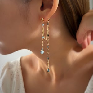 Op de afbeelding: Goudkleurige oorhangers met fijne kettingen en lichtblauwe kralen. De oorbellen hebben een kleine stud aan de bovenkant, met twee kettingen van verschillende lengtes. De kettingen zijn versierd met kleine en grote blauwe kralen, wat een waterval effect creëert.