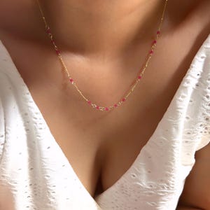 May include: Delicate gold necklace featuring small, vibrant red beads. The necklace is strung on a fine gold chain, creating a minimalist and elegant design. The necklace is shown against a white background.