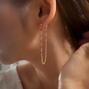 May include: Gold-toned earrings with a delicate design. One earring features a chain of small, vibrant pink beads, while the other has a fine chain with a dotted texture. The earrings are dangling, creating a graceful, elongated look.