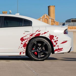 May include: White sedan with red splatter design on the side. The car has black wheels and red brake calipers. The vehicle is parked on a concrete surface under a clear blue sky.