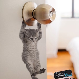 May include: A gray kitten decal hanging from a silver door knob. The decal is designed to look like a kitten hanging from the knob. The decal is made by StickerBrand and says "Made in USA".