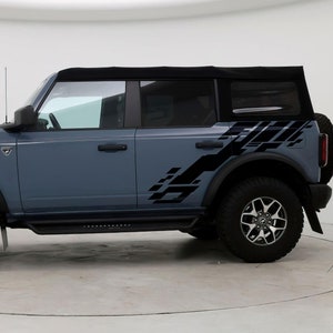 May include: A gray Ford Bronco with black side graphics. The vehicle has black wheels and a black soft top.