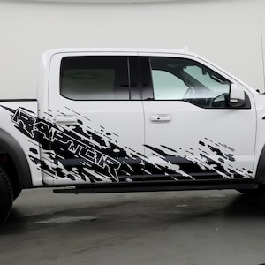 May include: White Ford F-150 Raptor pickup truck with black and white graphic decals on the side. The decals feature the word "RAPTOR" in a stylized font.