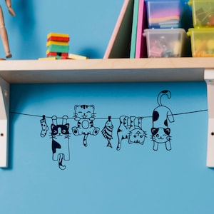 May include: A black and white wall decal of five cats hanging on a clothesline. The cats are holding socks, fish, and a ball of yarn.