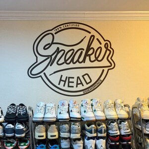 May include: A collection of sneakers displayed on a multi-tiered metal rack. The wall behind the rack features a black and white "Sneaker Head" logo with the text "100% Certified" and "@sneakerking15". The sneakers are in various colors and styles.