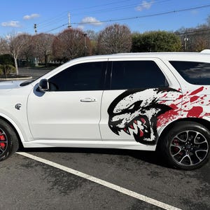 May include: White SUV with black wheels and red brake calipers. The side features a graphic of a snarling beast with red splatter accents. The vehicle is parked on a paved surface under a blue sky.