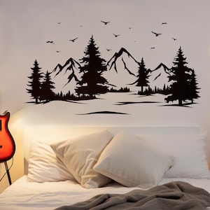 May include: Black and white silhouette wall decal of a mountain range with trees and birds. The decal is on a white wall above a bed with white bedding and pillows.