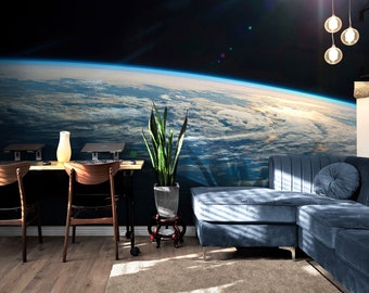 May include: A living room interior with a blue sectional sofa, a wooden table with two chairs, and a potted plant. The wall behind the sofa features a mural of Earth from space, showing the planet's blue oceans and white clouds.