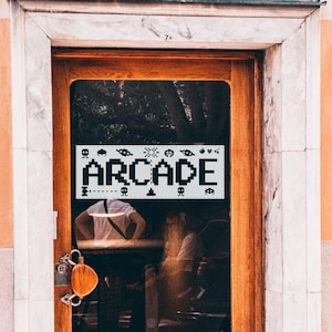 May include: A storefront window with a sign that says "ARCADE" in pixelated letters. The sign is decorated with pixelated images of video game characters and objects. The window is behind a wooden door with a metal handle.
