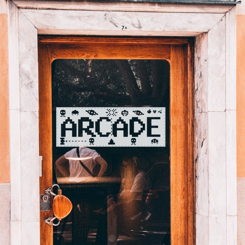 May include: A storefront window with a sign that says "ARCADE" in pixelated letters. The sign is decorated with pixelated images of video game characters and objects. The window is behind a wooden door with a metal handle.