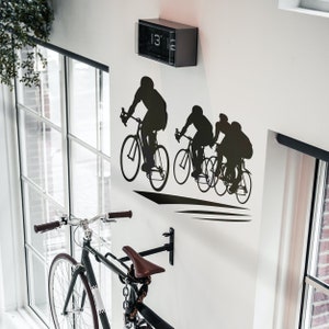 Puede incluir: Una silueta en blanco y negro de tres ciclistas montando bicicletas en una pared blanca. Los ciclistas est&aacute;n en l&iacute;nea, con el primer ciclista a la cabeza. Los ciclistas llevan cascos y tienen una mirada decidida en sus rostros. La pared est&aacute; decorada con un reloj negro con una esfera blanca y n&uacute;meros negros. El reloj marca las 13:15.