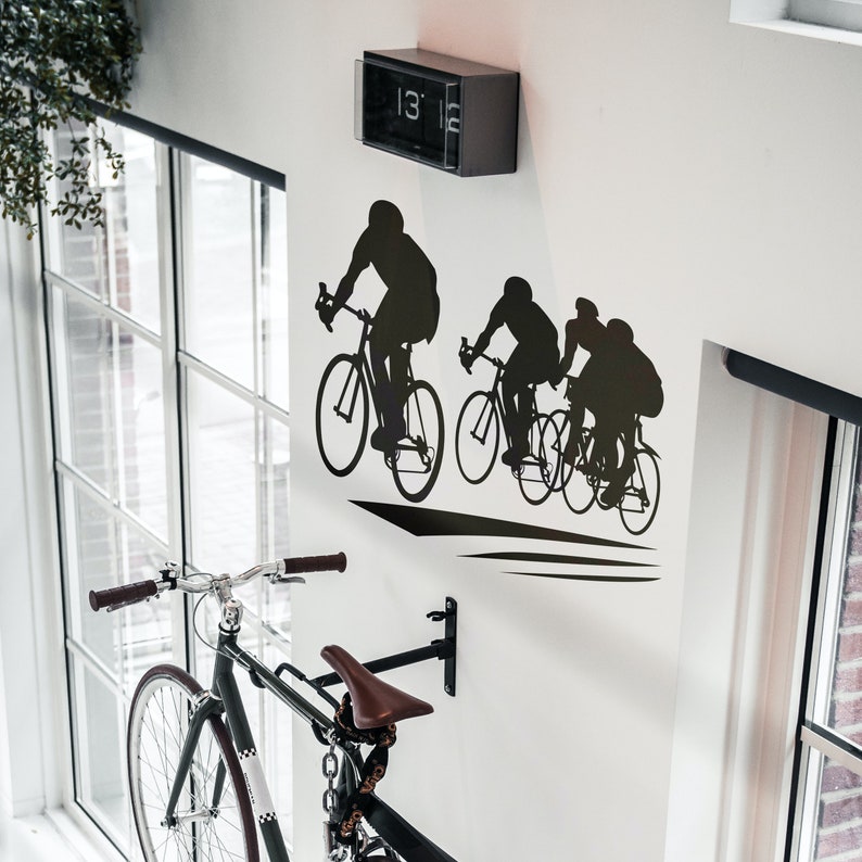 Puede incluir: Una silueta en blanco y negro de tres ciclistas montando bicicletas en una pared blanca. Los ciclistas est&aacute;n en l&iacute;nea, con el primer ciclista a la cabeza. Los ciclistas llevan cascos y tienen una mirada decidida en sus rostros. La pared est&aacute; decorada con un reloj negro con una esfera blanca y n&uacute;meros negros. El reloj marca las 13:15.