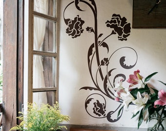 May include: A white wall with a black floral stencil design. The stencil features stylized flowers and leaves. There are white flowers in a vase in the foreground.