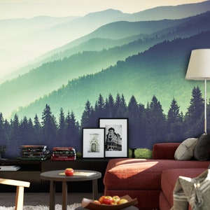 May include: A wall mural featuring a layered mountain landscape with green pine forests and misty peaks. The scene is set behind a red sofa, a dark side table with books, two small round tables with apples, and a floor lamp with a white shade.