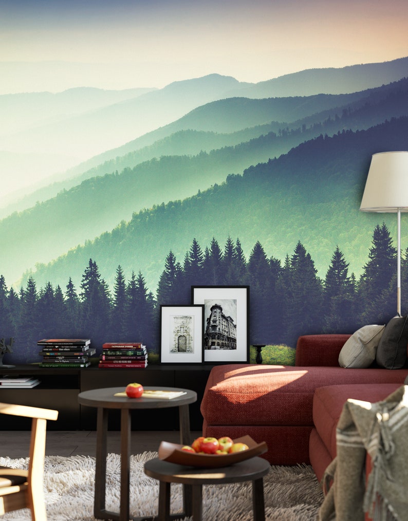 May include: A wall mural featuring a layered mountain landscape with green pine forests and misty peaks. The scene is set behind a red sofa, a dark side table with books, two small round tables with apples, and a floor lamp with a white shade.