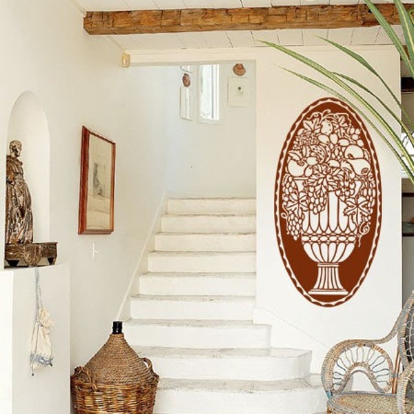 May include: A brown, decorative wall decal of a vase filled with fruit and grapes. The decal is in the shape of an oval and is placed on a white wall in a home interior setting.