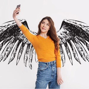 May include: A woman wearing a yellow sweater and blue jeans poses for a selfie with a smartphone in front of a white wall with a black and white drawing of angel wings.