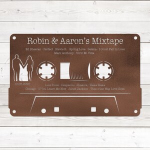May include: A brown metal cassette tape with a white outline and text that reads "Robin & Aaron's Mixtape". The cassette tape has a date of "10-05-2020" and a list of songs with artists, including "Ed Sheeran - Perfect", "Stevie B. - Spring Love", "Selena - I Could Fall In Love", "Marc Anthony - Vivir Mi Vida", "Luis Fonsi - Despacito", "Shakira - Waka Waka", "Chicago - If You Leave Me Now", and "Janet Jackson - That's the Way Love Goes".
