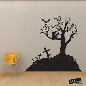 May include: Black and white Halloween themed wall decal featuring a spooky tree with a cat sitting on a branch, bats flying around, and three tombstones. The decal is on a grey wall with a yellow chair in the foreground.