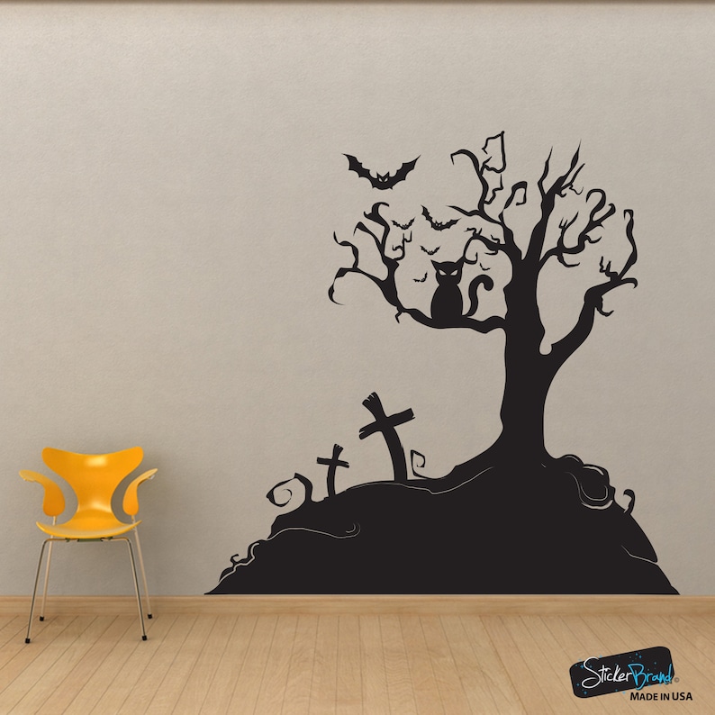 May include: Black and white Halloween themed wall decal featuring a spooky tree with a cat sitting on a branch, bats flying around, and three tombstones. The decal is on a grey wall with a yellow chair in the foreground.
