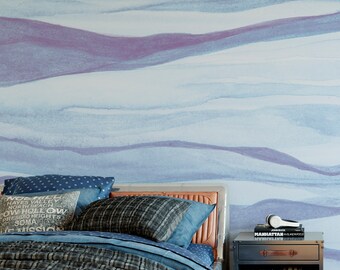 May include: A bedroom scene featuring a bed with blue and gray patterned pillows and bedding, set against a wall with a horizontal wavy watercolor mural in shades of light blue and purple. A metal nightstand holds books and white headphones.