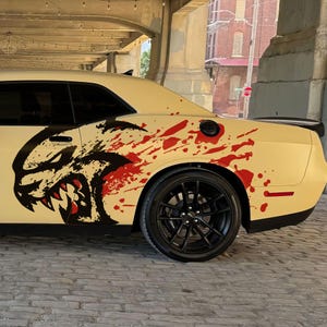 May include: Cream-colored sports car with a black and red graphic design on the side. The design features a snarling face and red splatter accents. The car has black wheels and is parked on a cobblestone street.