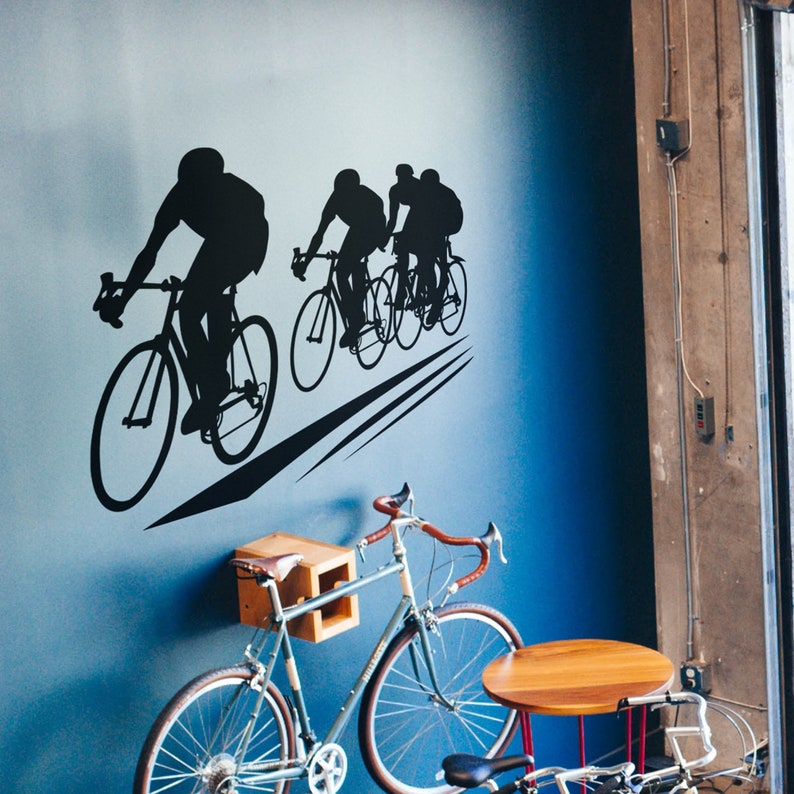 Puede incluir: Una calcoman&iacute;a de pared en silueta negra y blanca de tres ciclistas montando bicicletas. La calcoman&iacute;a est&aacute; en una pared azul con una bicicleta apoyada contra la pared.