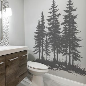 May include: A gray wall decal of several tall pine trees in a bathroom setting. The trees are silhouetted against a light gray wall, with a white toilet and wooden vanity in the foreground. The floor is a light gray marble.