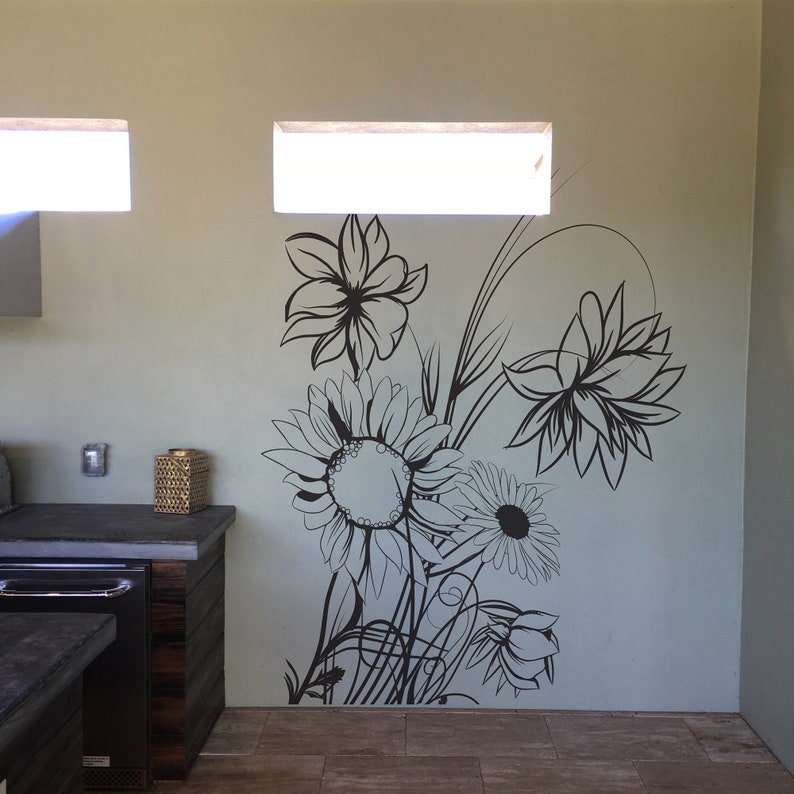 Op de afbeelding: Een zwart-witte muursticker met een bloemenontwerp met zonnebloemen, lelies en andere bloemen. De sticker zit op een lichtblauwe muur.