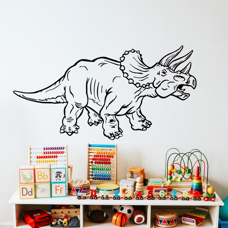 K&ouml;nnte beinhalten: Schwarz-wei&szlig;-Zeichnung eines Triceratops-Dinosauriers an einer wei&szlig;en Wand. Der Dinosaurier ist nach rechts gerichtet und hat einen gro&szlig;en Nackenschild und drei H&ouml;rner. Der Dinosaurier befindet sich in einem Spielzimmer mit Spielzeug in einem Regal darunter.
