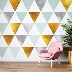 May include: A wall covered in wallpaper featuring a repeating geometric pattern of triangles in metallic gold, silver, and pale mint green colors. A light pink upholstered armchair with light-colored wooden legs sits in front of the wall.