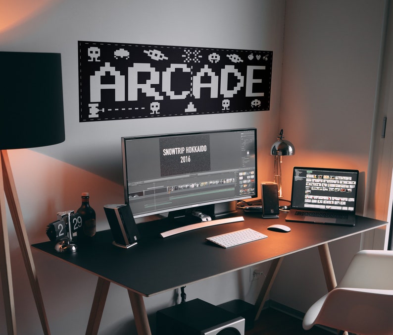 May include: A black desk with a white keyboard, a black computer monitor, a black laptop, a black lamp, and a black speaker. The monitor displays a video editing program with the text "SNOWTRIP HOKKAIDO 2016". The wall behind the desk has a black and white banner with the word "ARCADE" in pixelated letters.