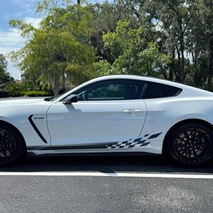 May include: A white sports car with black wheels and a checkered flag design on the side. The car is parked on an asphalt surface with trees in the background. The car has a sleek, aerodynamic design.
