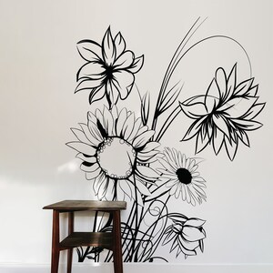 Op de afbeelding: Zwart-witte lijntekening van een boeket bloemen op een witte muur. De bloemen omvatten zonnebloemen, madeliefjes en andere bloemmotieven. Een kleine houten tafel met twee planken staat op de voorgrond.