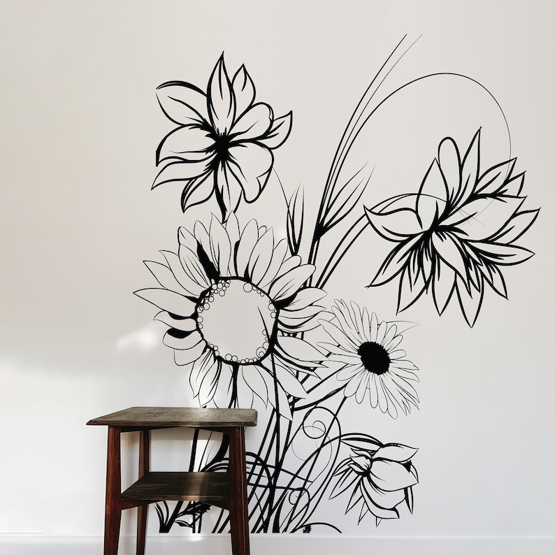 Op de afbeelding: Zwart-witte lijntekening van een boeket bloemen op een witte muur. De bloemen omvatten zonnebloemen, madeliefjes en andere bloemmotieven. Een kleine houten tafel met twee planken staat op de voorgrond.