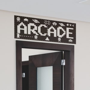 May include: A black and white wall decal with the word "ARCADE" in pixelated letters. The decal is surrounded by small pixelated images of video game characters and objects. The decal is above a dark brown wooden door.