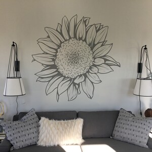 Puede incluir: Un sof&aacute; gris con dos almohadas blancas y una almohada de estampado gris y blanco. El sof&aacute; est&aacute; frente a una pared con un gran adhesivo de girasol gris. Hay dos apliques de pared negros con pantallas blancas a cada lado del sof&aacute;.