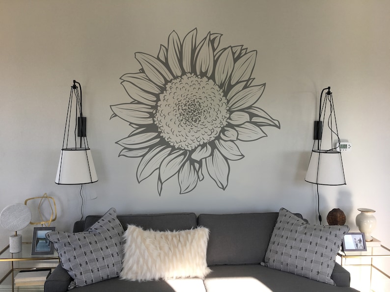 Puede incluir: Un sof&aacute; gris con dos almohadas blancas y una almohada de estampado gris y blanco. El sof&aacute; est&aacute; frente a una pared con un gran adhesivo de girasol gris. Hay dos apliques de pared negros con pantallas blancas a cada lado del sof&aacute;.